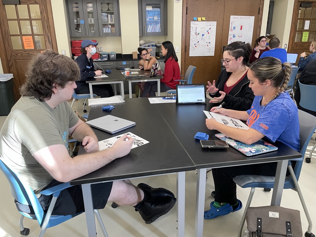 5DC4108B-A2A8-4E34-ABE0-26A55138E684_1_105_c Students collaborate in a classroom at the University of Florida College of Education. Several students are seated around black tables with blue chairs, working on papers and laptops. The room has storage cabinets on the walls and educational posters displayed. One student wears a blue UF shirt, and the setting appears to be a typical UF Teach program working session. Some students are engaged in discussion while others are focused on individual work. The environment suggests an active learning space within an educational setting